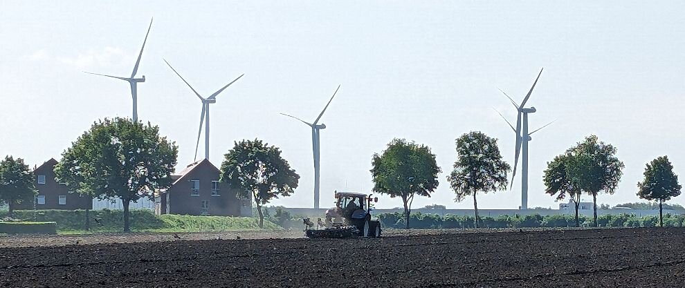 Foto eines Schleppers bei der Feldarbeit. Im Hintergrund Windräder